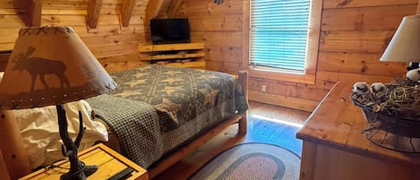 A cozy cabin room with a bed, a desk, a lamp, a television, and a window with blinds.