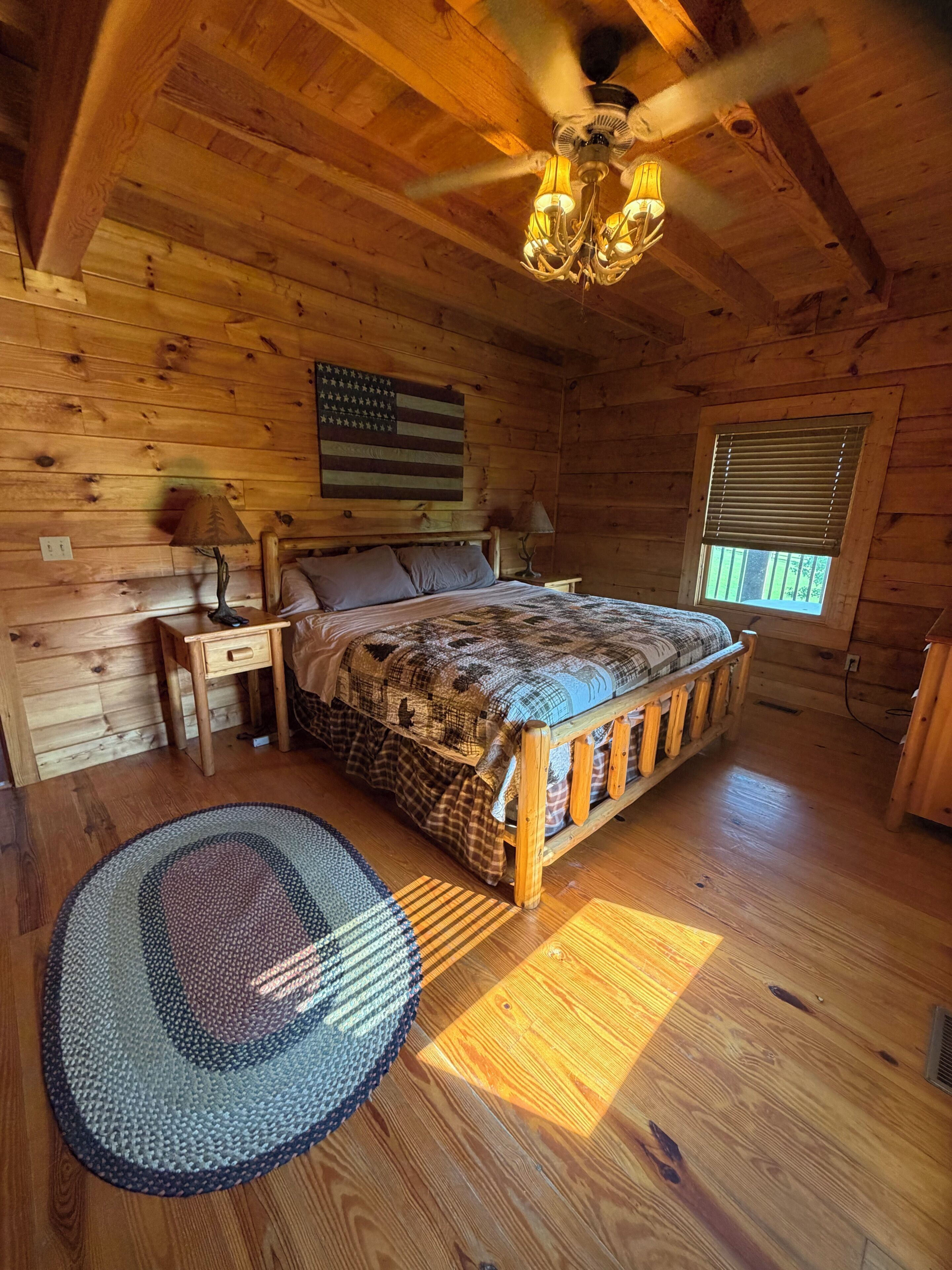 A wooden cabin bedroom with a bed, two nightstands, a ceiling fan, and a window with blinds.