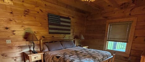 Un dormitorio de cabaña de madera con una cama, dos mesitas de noche, un ventilador de techo y una ventana con persianas.