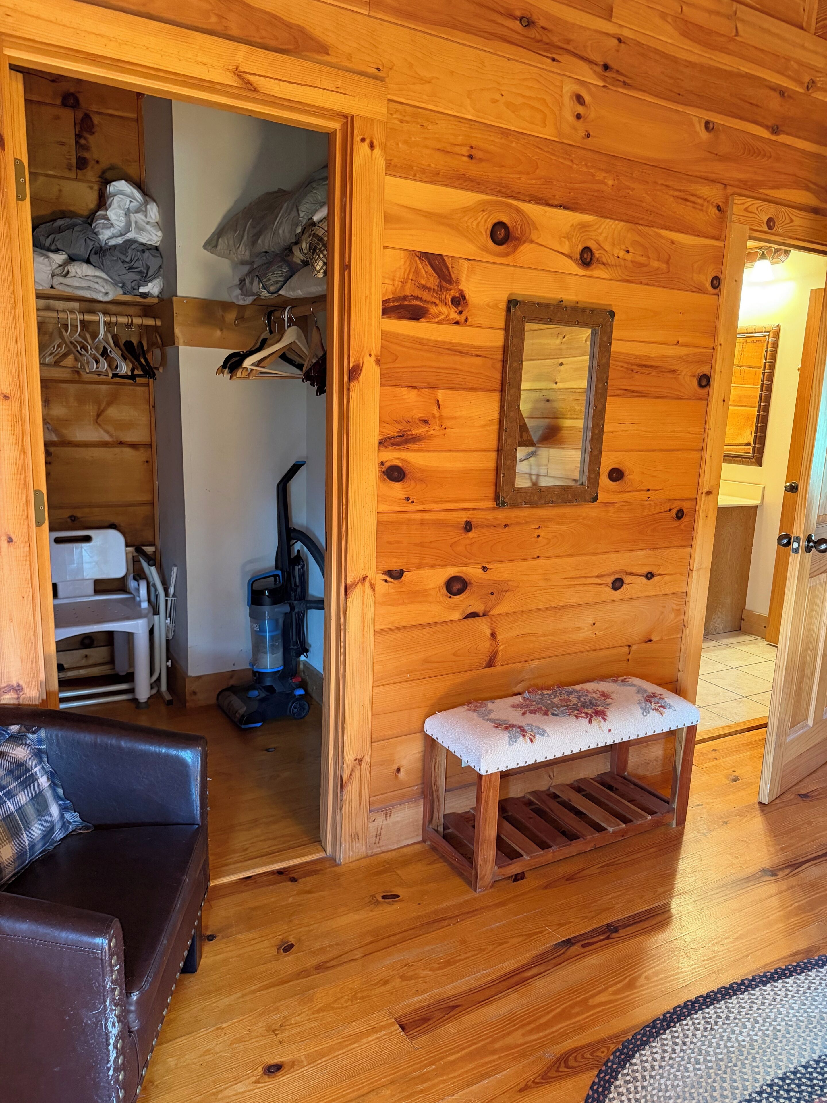 A wooden interior with a bench, a closet with hanging clothes, and a vacuum cleaner.