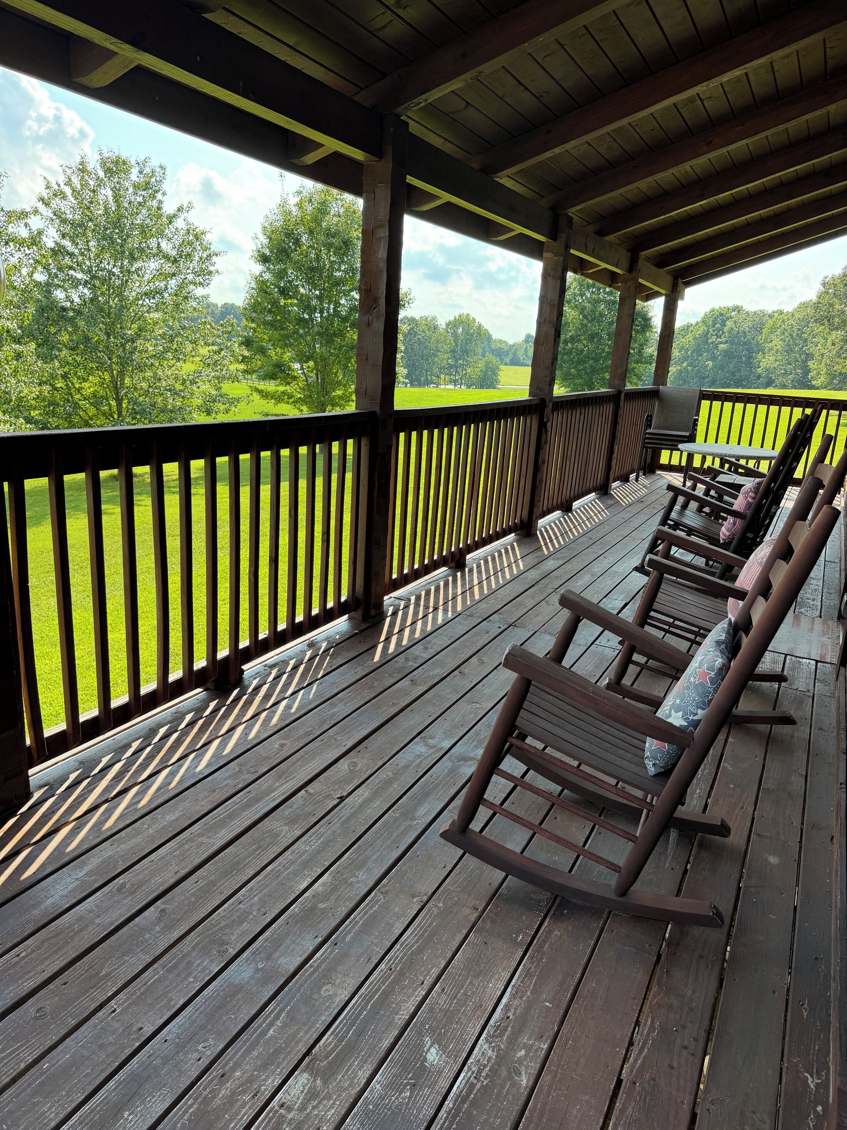 Une terrasse en bois offrant une vue sur un champ verdoyant et des arbres. On y trouve des chaises et une table en bois.