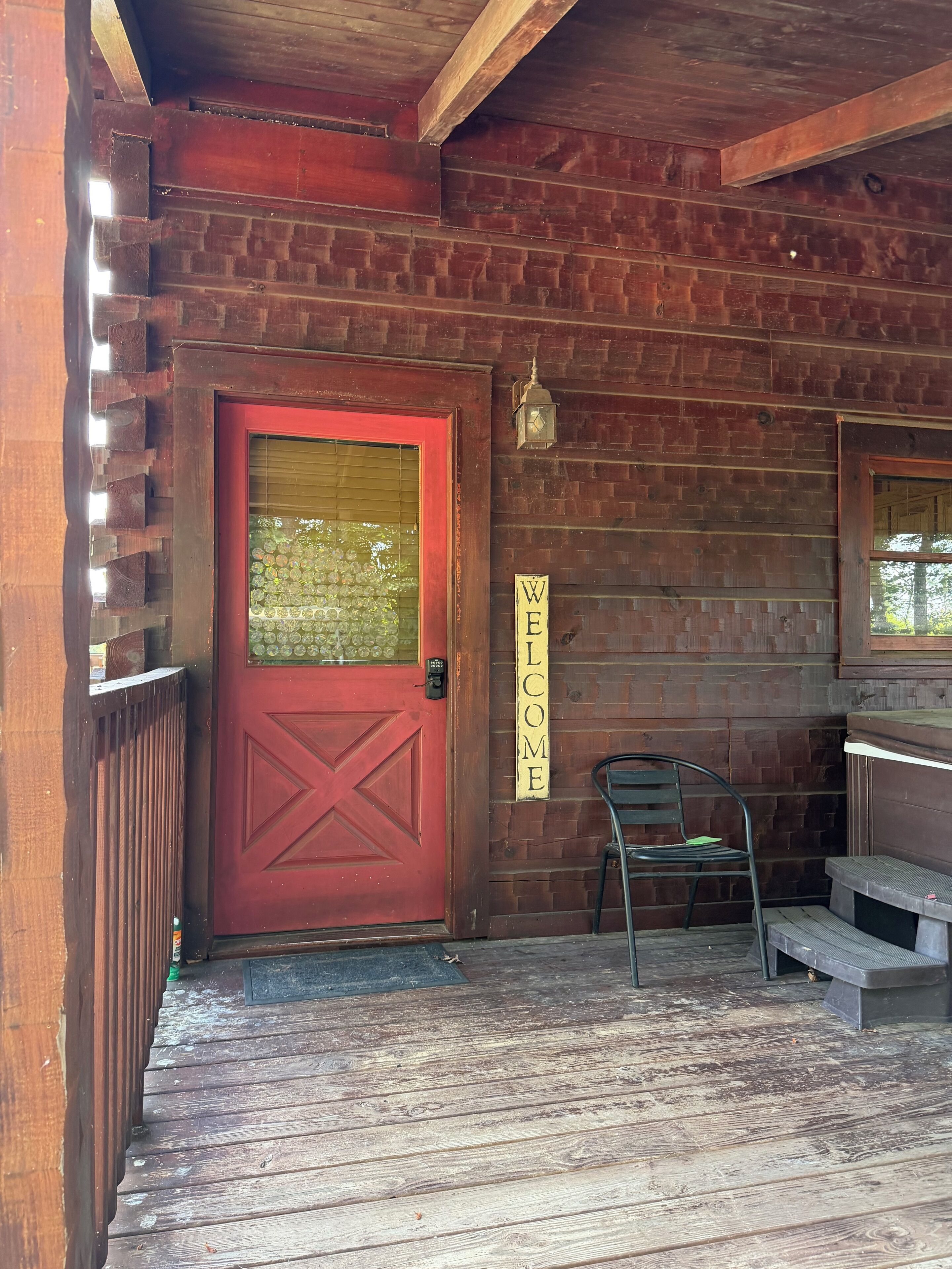 A wooden door with a red frame and a welcome sign, a bench, and a lantern on a porch with wooden railings.