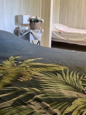 A bed with a green leaf-patterned bedspread, a bedside table with a basket of white flowers, and a white mattress with a floral cover.