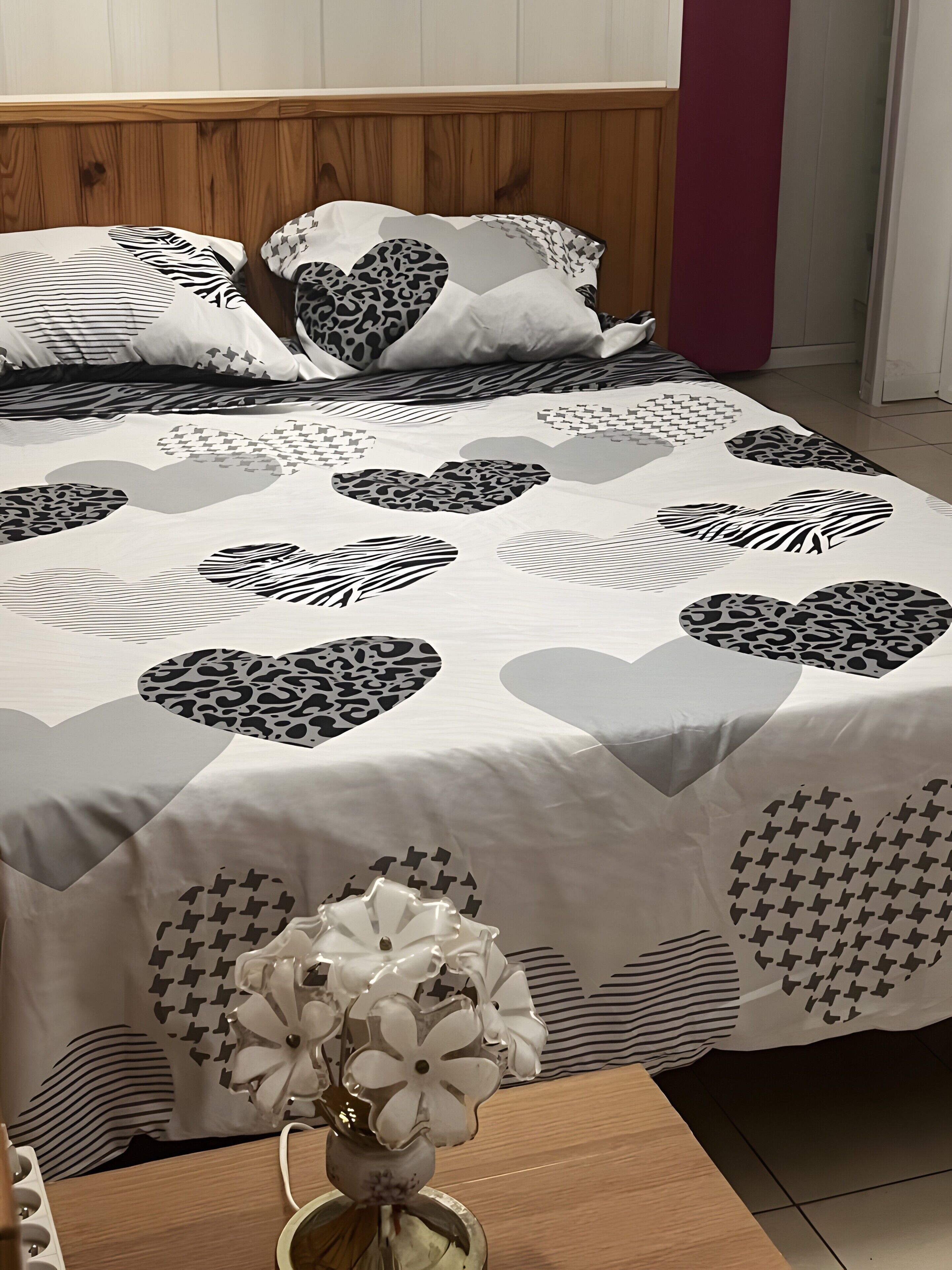 Uma cama com colcha estampada com corações e desenhos abstratos, uma mesinha de cabeceira com um vaso de flores e uma cabeceira de madeira.