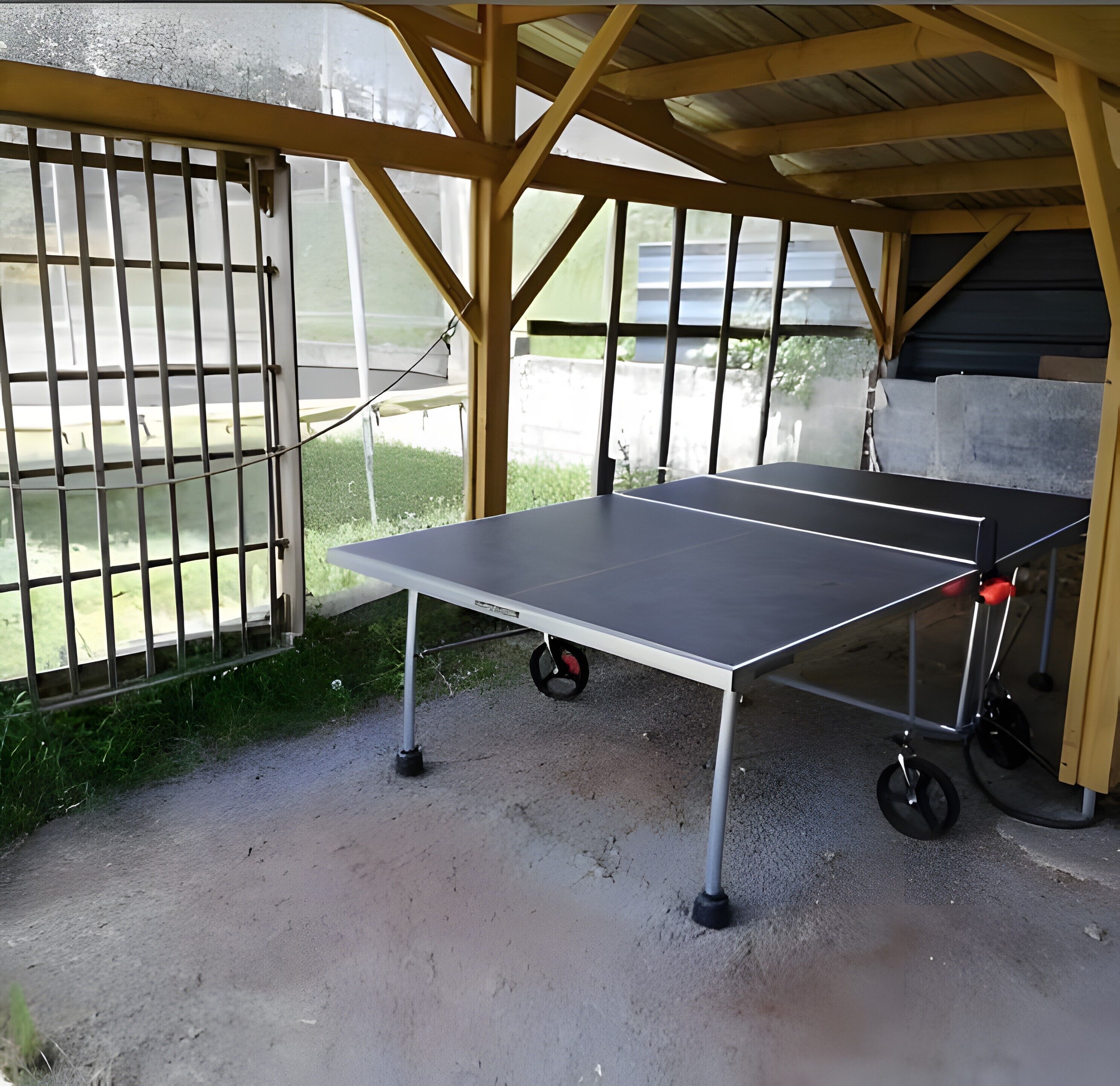 Una mesa de ping pong con ruedas, montada al aire libre bajo un techo de madera con barandilla de metal.