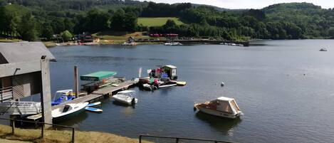 A lakeside dock with boats, a small building, and a forested hill in the background.