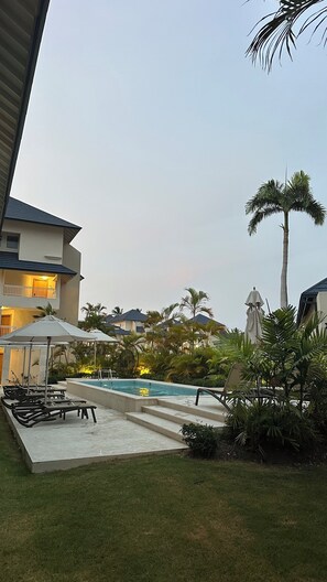 A pool area with lounge chairs and umbrellas, surrounded by palm trees and a building with balconies. -  XANADU CASA DE LA PLAYA (Puerto Plata)