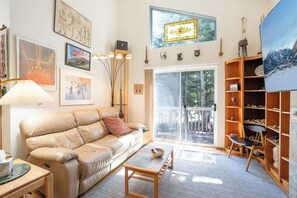 Ein Wohnzimmer mit Ledersofa, Couchtisch, Bücherregal und einem großen Fenster mit Blick auf Bäume.