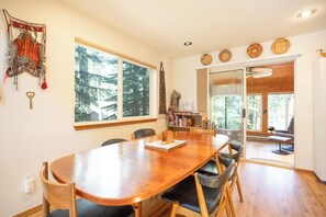 Ein Holztisch mit Stühlen, ein Bücherregal und Blick auf Bäume durch große Fenster.