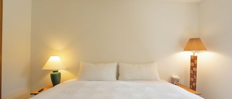 Una cama bien hecha con ropa blanca, dos lámparas de noche y una mesita de noche pequeña con un florero y una caja de pañuelos.