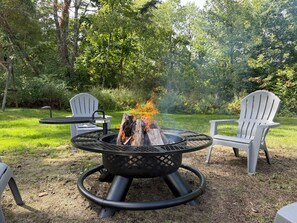 A circular outdoor fire pit with a metal grate, surrounded by two Adirondack chairs and a small table. - 3-bedroom house in peaceful, rural hideaway (Schenectady)
