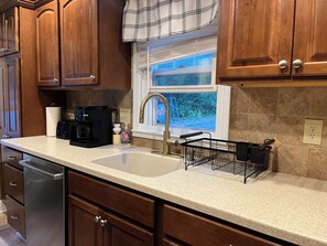 A kitchen with wooden cabinets, a stainless steel dishwasher, a black coffee maker, and a window with a view of greenery. - 3-bedroom house in peaceful, rural hideaway (Schenectady)