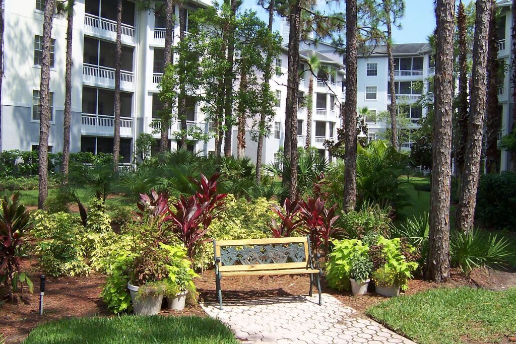 Un immeuble à plusieurs étages doté de balcons, entouré d’un jardin aménagé avec différentes espèces de plantes et agrémenté d’un banc central.