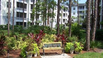 Un immeuble à plusieurs étages doté de balcons, entouré d’un jardin aménagé avec différentes espèces de plantes et agrémenté d’un banc central.