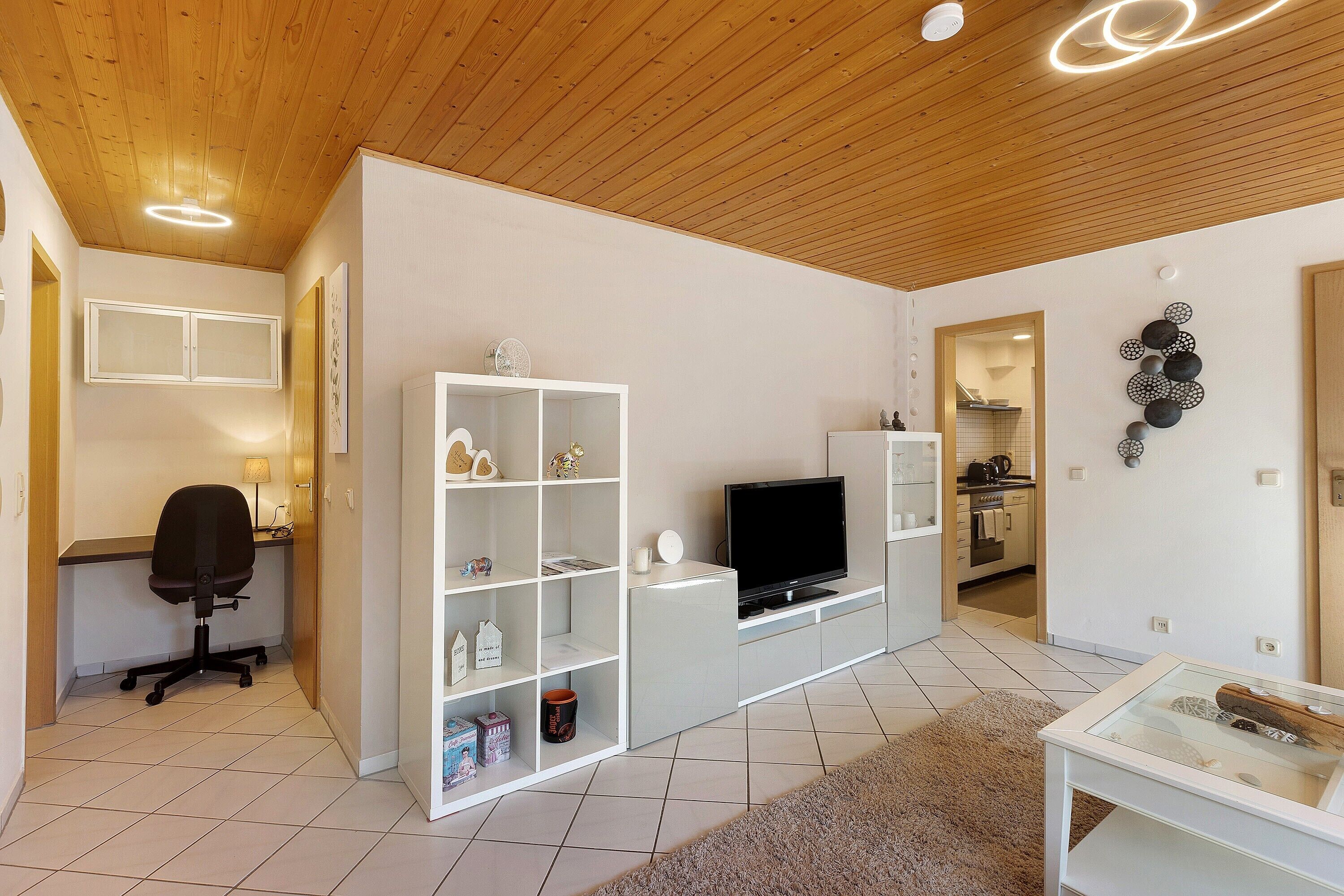 Ein modernes Wohnzimmer mit Fernseher, einem weißen Regalschrank und einem Schreibtisch mit Stuhl.