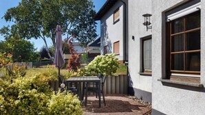 Eine Terrasse mit Tisch und Stühlen, umgeben von Grünpflanzen und einem weißen Gebäude mit Fenstern.