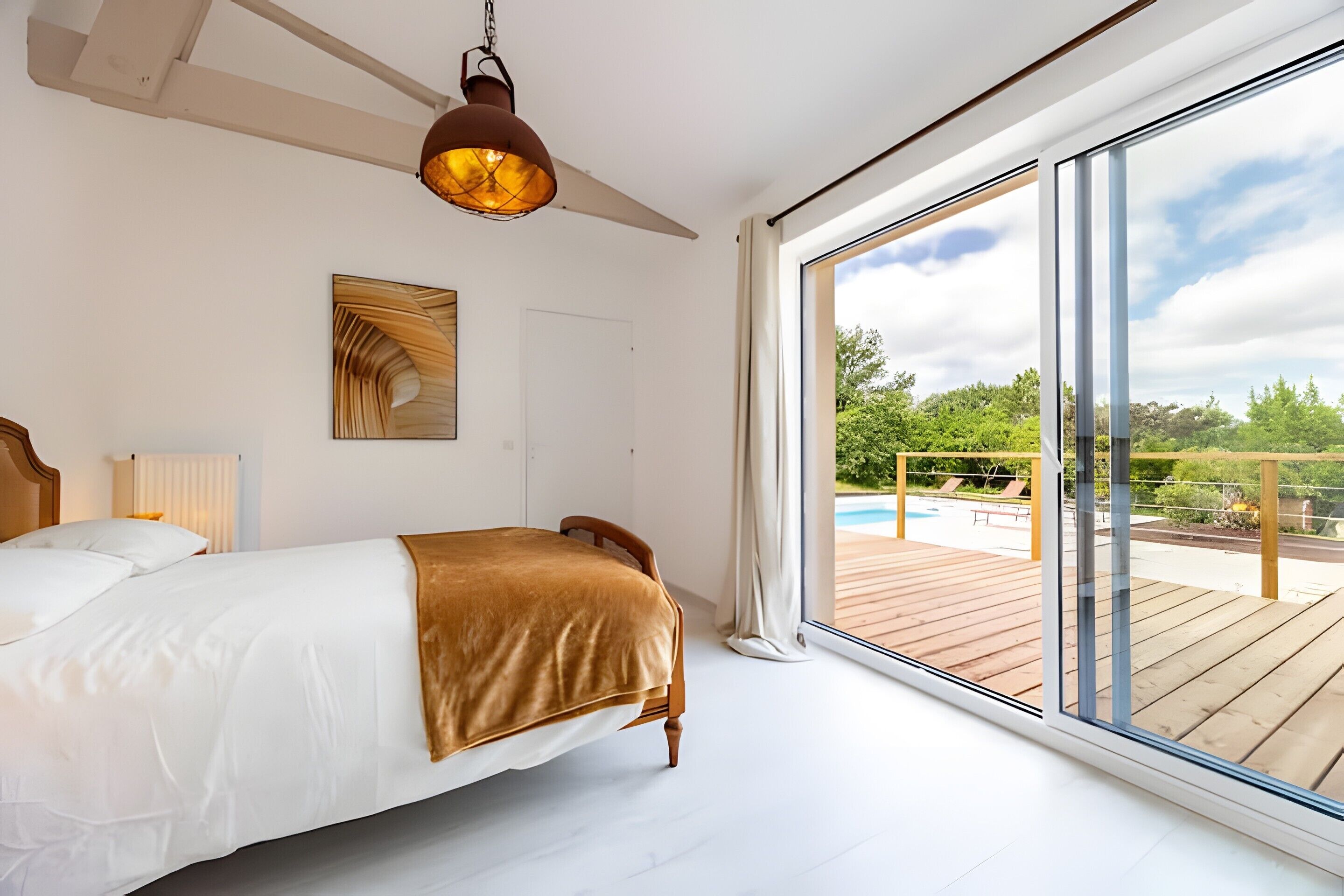 A bedroom with a large bed, a ceiling fan, a painting, and a view of an outdoor pool area.
