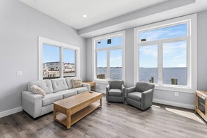 Un salon moderne avec un canapé gris, une table basse en bois et deux fauteuils gris. De grandes fenêtres offrent une vue sur l’eau et les bâtiments.