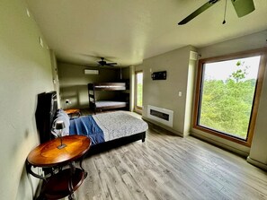 Quarto com cama beliche, bateria, ventilador de teto e uma grande janela com vista para o verde.