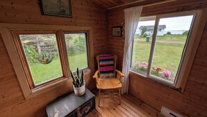A cabin with wooden walls, a bench, a colorful chair, and a window overlooking a grassy yard.