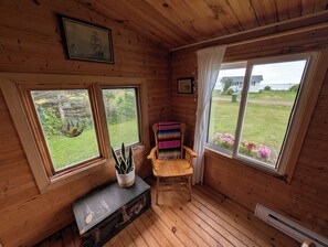 A cabin with wooden walls, a bench, a colorful chair, and a window overlooking a grassy yard.