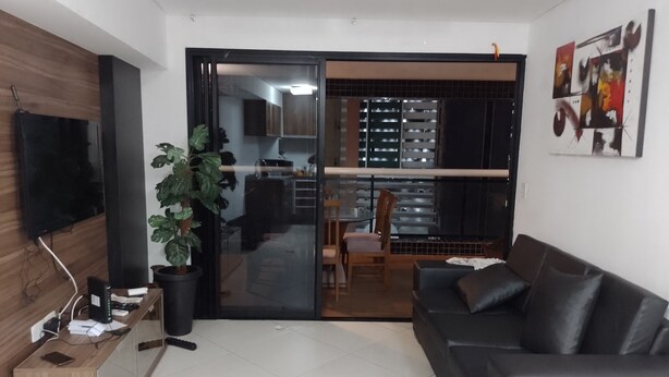A modern living room with a black leather sofa, a wooden TV stand, a potted plant, and a sliding glass door leading to a kitchen and dining area. - Landscape Apartment in Fortaleza Gold (Fortaleza)