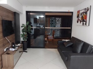 A modern living room with a black leather sofa, a wooden TV stand, a potted plant, and a sliding glass door leading to a kitchen and dining area. - Landscape Apartment at Fortaleza Gold (Fortaleza)