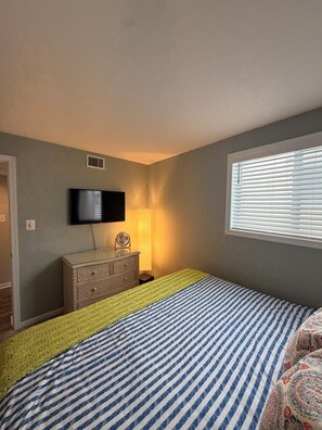 A bedroom with a bed, a television mounted on the wall, a bedside table with a lamp, and a window with blinds. - 2-bedroom condo in lovely Wildwood Crest -1 block to Beach-Bikes-Ice cream! (Wildwood Crest)