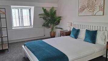 A bedroom with a bed, a nightstand, a bedside table, a hanging plant, a radiator, a window with blinds, and a coat rack.