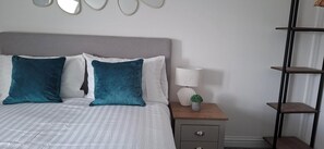 Ein Bett mit grauem Kopfteil, zwei türkisfarbenen Kissen und einem weißen Kissen. Ein Nachttisch mit Lampe, einer kleinen Pflanze und einem Holzregal.