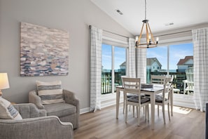 A modern living room with a dining area, large windows, and a view of the outside. - Top-Floor Ocean Views | Pool + Beach Access (Emerald Isle)