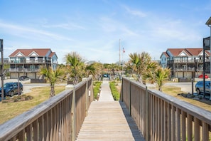 A wooden boardwalk leading to a residential area with houses, a flag, and parked cars. - Top-Floor Ocean Views | Pool + Beach Access (Emerald Isle)
