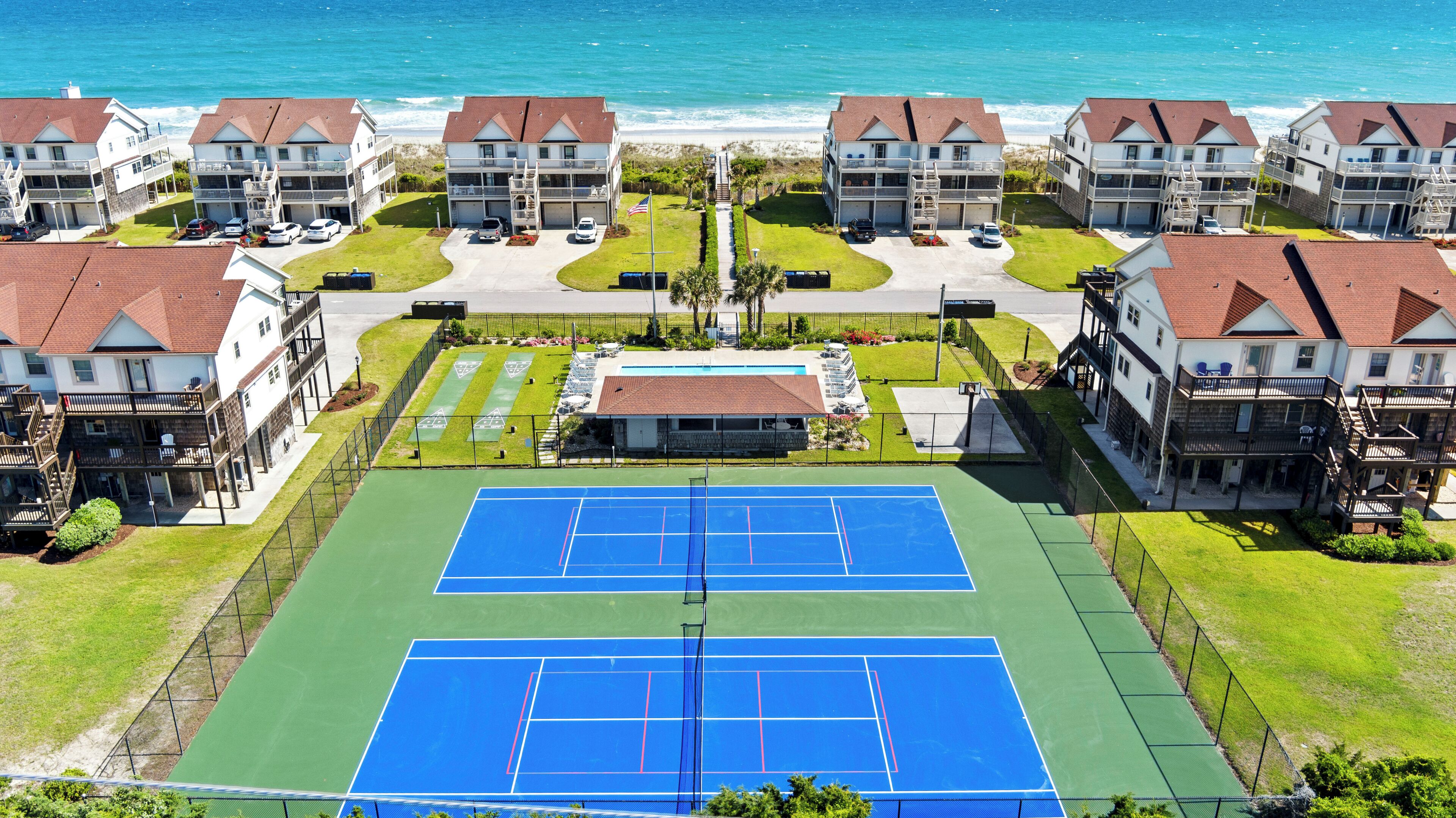 Un complexe résidentiel situé en bord de mer, composé de plusieurs maisons à deux étages, d’un court de tennis central et offrant une vue sur la plage.