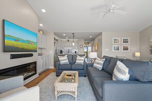 A modern living room with a flat-screen TV, a fireplace, a ceiling fan, and a kitchen in the background. - Top-Floor Ocean Views | Pool + Beach Access (Emerald Isle)