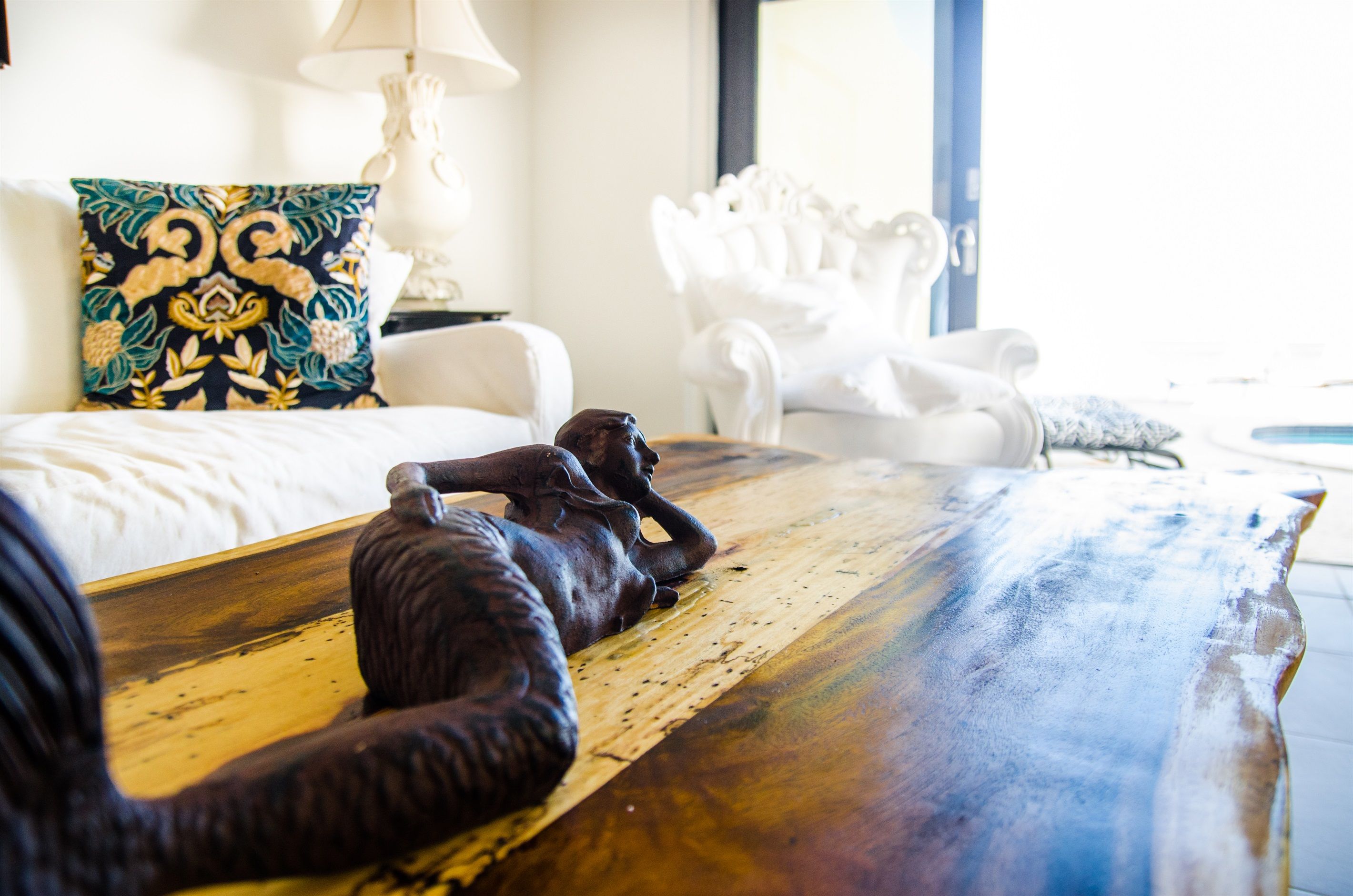 A bed with a floral pillow, a white armchair, and a wooden table with a sculpture.