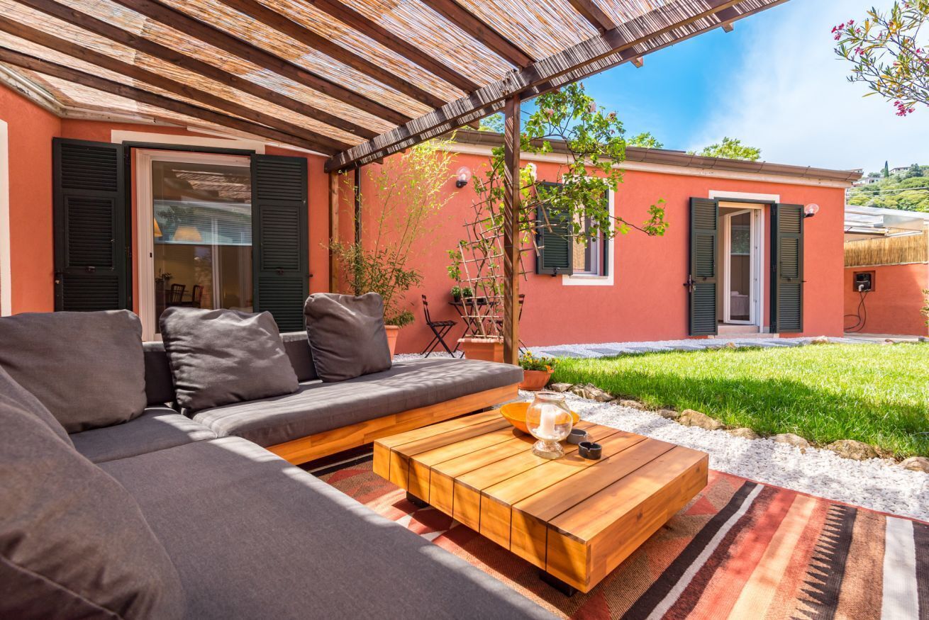 Un'accogliente area salotto all'aperto con divano e tavolino in legno, una pergola sopra e un piccolo giardino con un albero e un edificio sullo sfondo.