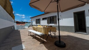 A patio with a table and bench, a large umbrella, and a building with windows and a door. - Casa Turalves - By WeHostMadeira (Porto Santo)