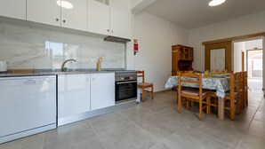 A modern kitchen with white cabinets, a built-in oven, and a dining area with wooden chairs and a table. - Casa Turalves - By WeHostMadeira (Porto Santo)