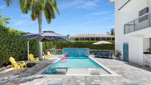 A swimming pool with lounge chairs and umbrellas, surrounded by a hedge and a building. - Brand new, walking distance to Sombrero Beach! 4 Bed/4.5 Bath outdoor kitchen. (Marathon)