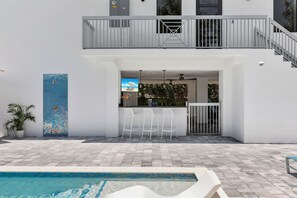Ein moderner Poolbereich im Freien mit Bar und Sitzgelegenheiten, einer überdachten Terrasse und einer Treppe, die zu einer erhöhten Ebene führt.