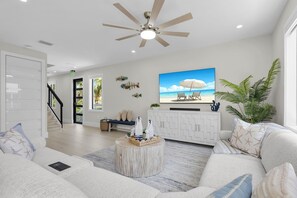 Ein modernes Wohnzimmer mit Deckenventilator, einem Fernseher, der eine Strandansicht zeigt, einem weißen Schrank und einer großen Pflanze.