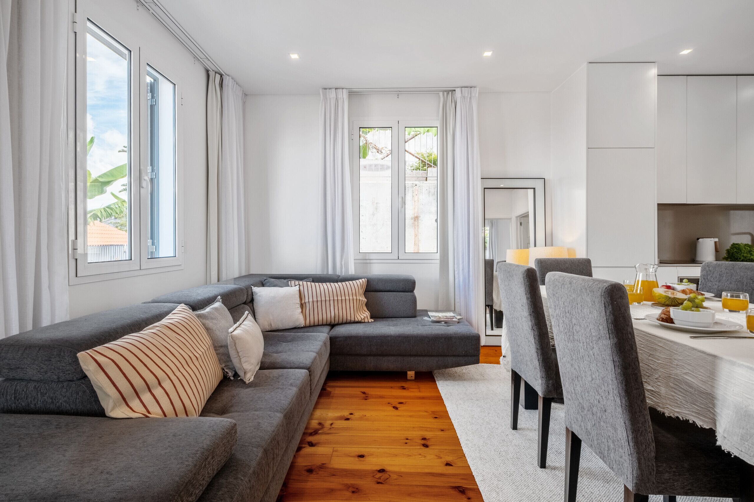 A modern living room with a grey sectional sofa, a dining table with chairs, and large windows with curtains.
