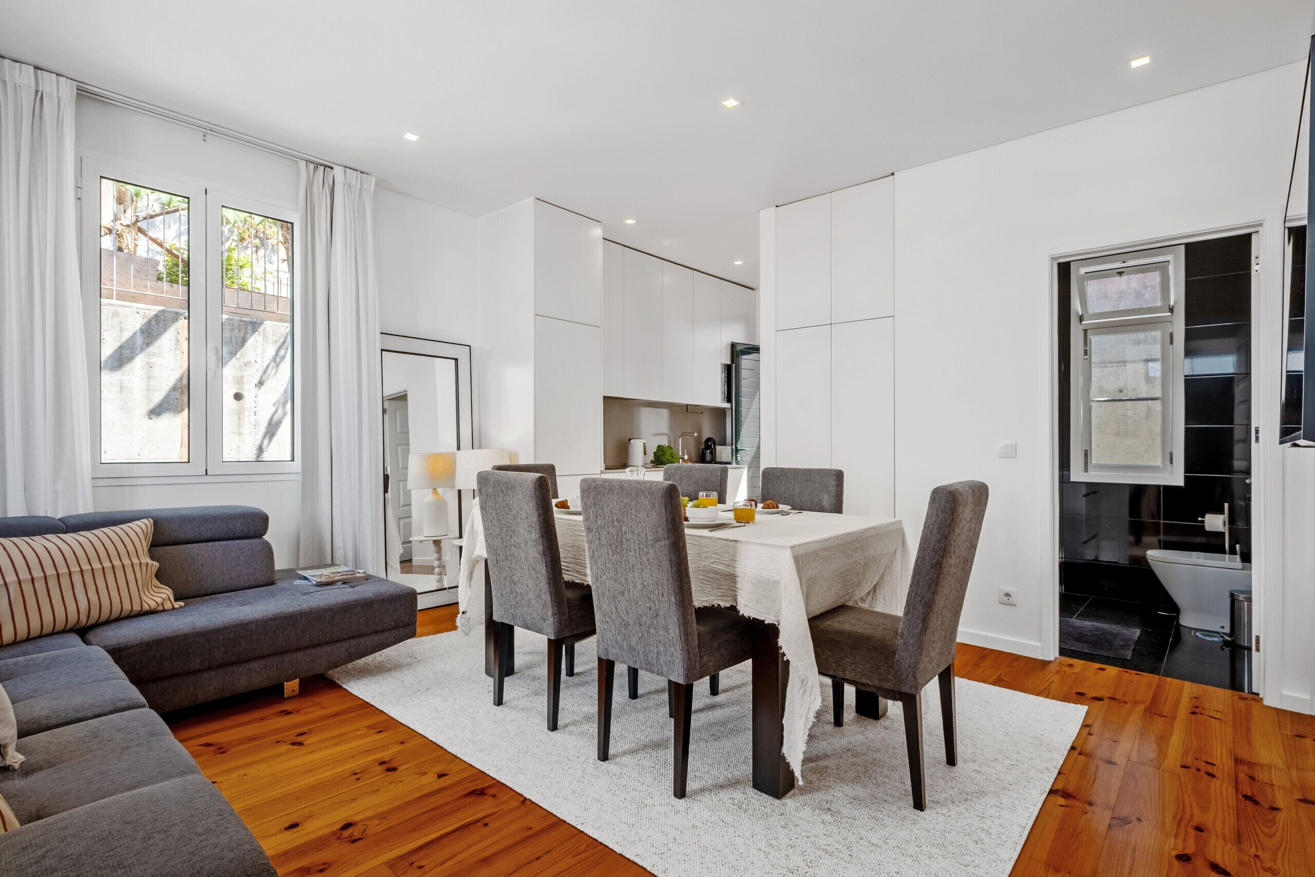 A modern living room with a grey sofa, a dining table with chairs, and a kitchen area in the background.
