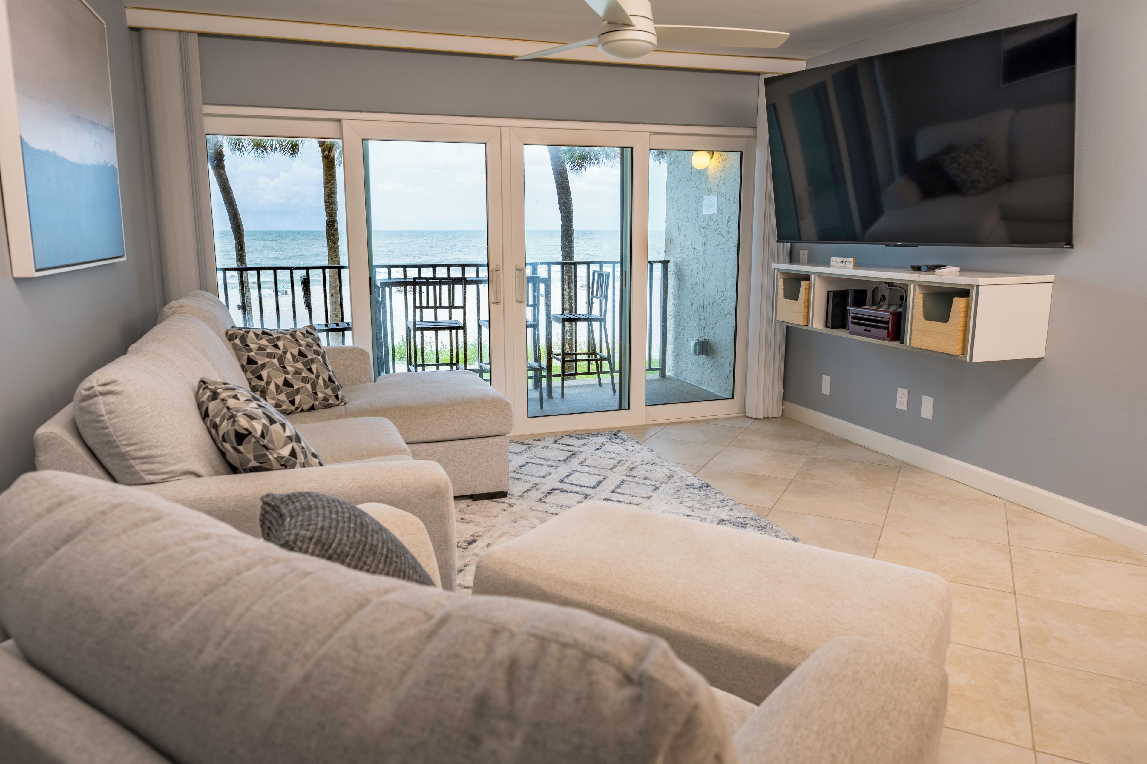 A living room with a large flat-screen TV, a sofa, and a view of the ocean through glass doors.