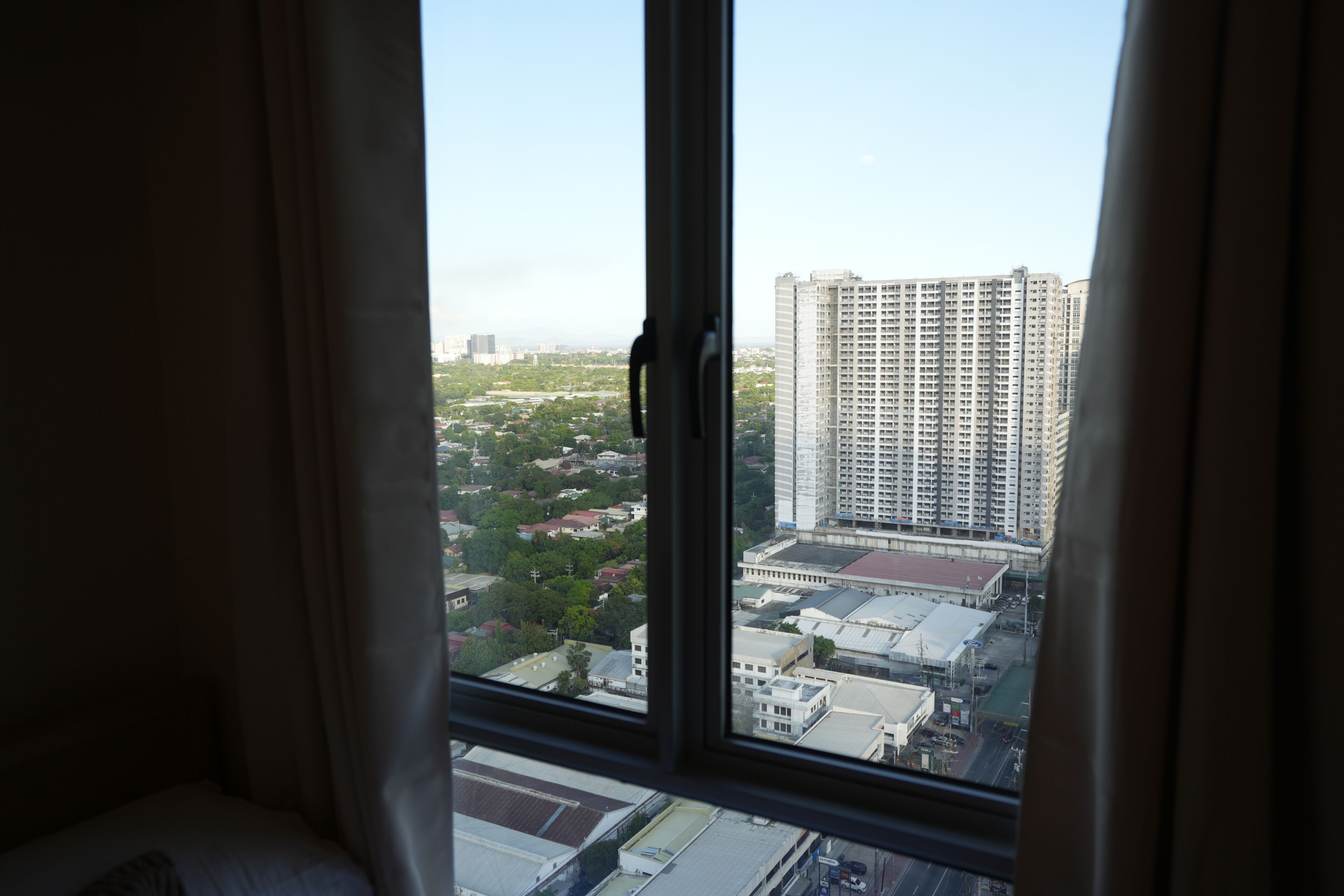 Una vista attraverso una finestra con tende, che mostra un panorama urbano con grattacieli e un mix di edifici residenziali e commerciali.