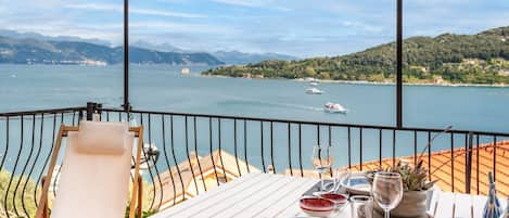 Una terraza con una mesa lista para comer, con vista a un cuerpo de agua donde se ven barcos y colinas a lo lejos.