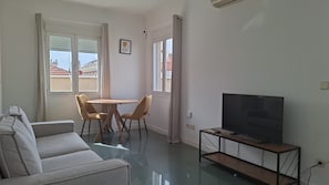 A living room with a sofa, a TV on a stand, a dining table with chairs, and large windows with curtains. - Ideal Accommodations in Madrid – Calle Cicerón with a Terrace (MADRID centro)