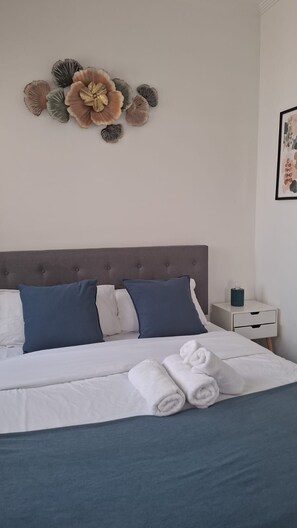 Ein ordentlich bezogenes Bett mit weißer und blauer Bettwäsche, einem gepolsterten Kopfteil und einem dekorativen Wandbild darüber.