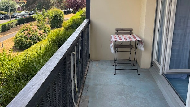 En balkong med ett litet bord och en stol, utsikt över en väg och grönska, och en byggnad i bakgrunden.