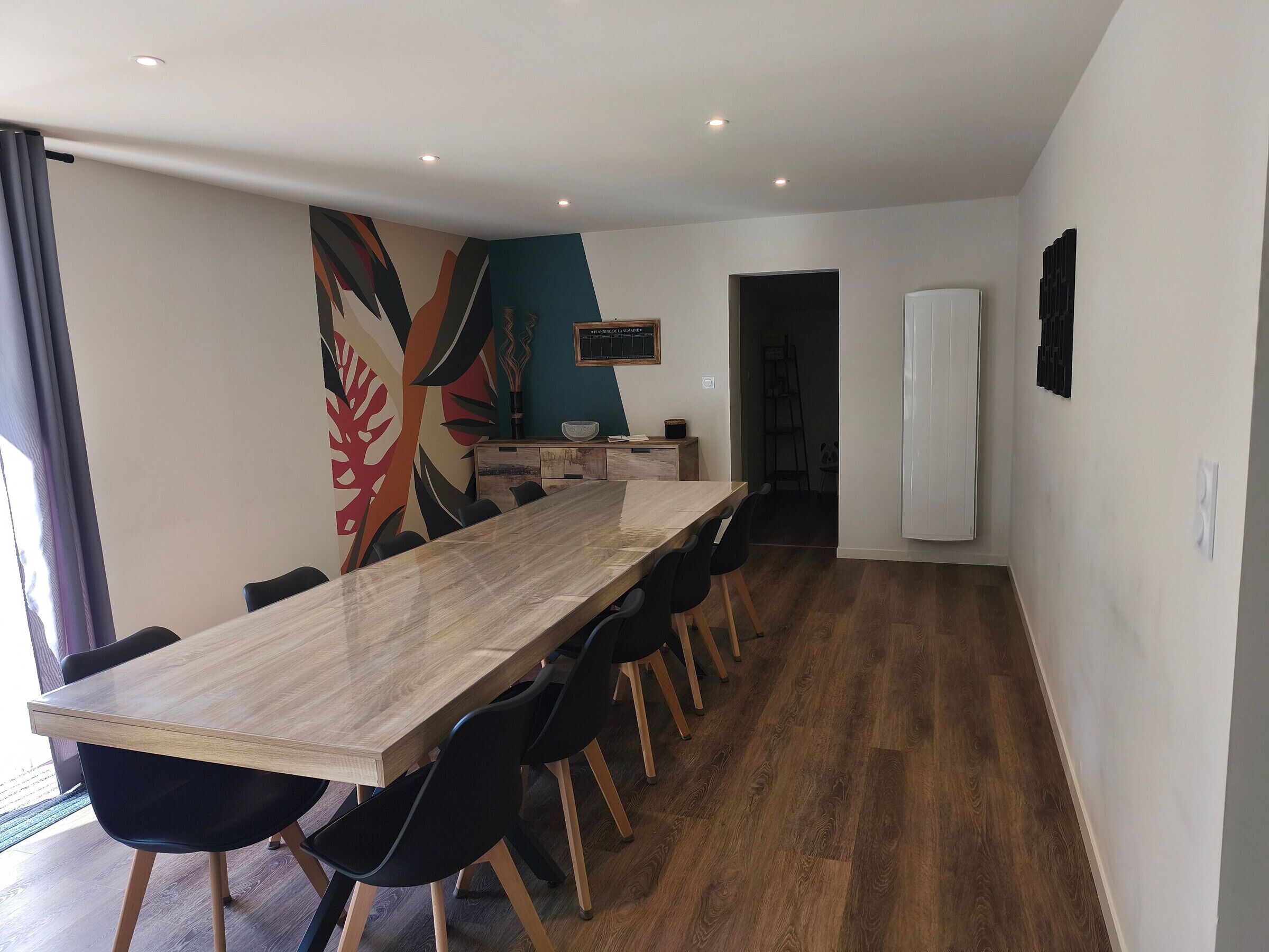 A modern dining area with a long wooden table, black chairs, and a colorful abstract wall mural.
